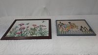 Two framed embroidery artworks laid side by side on white quilted surface, showing floral and cottage scenes