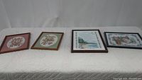 Four framed hand embroidered needlepoint artworks laid flat on a light quilted surface showing full lot.