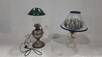 Two vintage oil lamps, one electrified Rayo kerosene oil lamp with green metal shade and one clear glass lamp with floral fabric shade, displayed together.