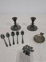 Top view photo showing two silver candle holders, five silver spoons, a pewter pineapple trinket dish, and a pewter tree paperweight laid on a white surface.