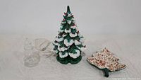Vintage green ceramic Christmas tree with attached power cord and base, next to clear glass bulb and small ceramic dish on cloth surface