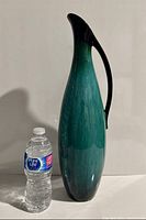 Front view of tall teal vase with black handle next to water bottle for scale
