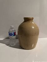 Front view of large beige stoneware pot standing upright next to water bottle for scale, showing smooth glazed surface with speckles and incised decoration.