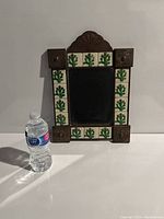 Front view of a rectangular mirror framed by ceramic tiles with green cactus patterns and brown wooden decorative blocks, shown next to a water bottle for size reference.