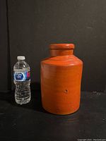 Front view of an orange cylindrical stoneware pot alongside a water bottle for size reference.