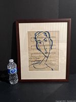 Framed artwork with blue crayon figure of a woman on printed musical sheet, accompanied by water bottle for scale.