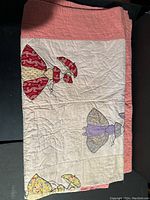 Quilt folded showing embroidered women in bonnets and colorful dresses holding umbrellas. Light beige fabric with pink border.