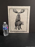 Photo showing entire print with a water bottle for scale, depicting two moose in black ink on beige paper.