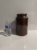 Stoneware pot standing upright next to a water bottle for size reference, showing brown glazed surface and cylindrical shape.