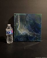 Full view of 12x12 acrylic painting next to water bottle for scale, showing square canvas artwork with dark moody colors and fluid style.