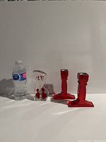 Image shows two red glass candlesticks and one small clear glass figurine with red decoration alongside a water bottle for size reference.