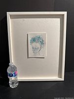 Full view of the framed watercolour portrait with a water bottle for scale, showing white frame and large matting around the small artwork.