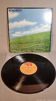 Front view of the Blind Faith vinyl LP record partially out of its album cover, showing the artwork of green field and cloudy blue sky on the cover.
