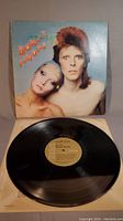 Photo of the front cover of David Bowie Pin Ups album with record partially in view. Cover shows a portrait of David Bowie and a woman with some visible wear and touch up.
