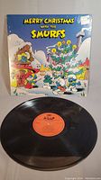 Photo showing the front cover of Merry Christmas With The Smurfs with several Smurfs cartoon characters in snowy Christmas scene, alongside the black vinyl record with visible grooves and center label partially visible.