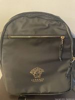 Front view of the black backpack showing the gold zipper and Versace Parfums logo with Medusa head.