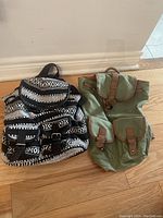 Photo showing two backpacks on a wooden floor against a white wall. One backpack is black and white with geometric patterns and multiple buckled pockets. The other backpack is green canvas with brown straps and buckles.