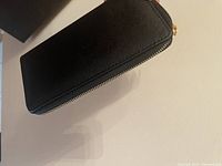 Closed black zip-around wallet showing textured exterior and gold-tone zipper pull.