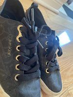 Close-up side view of black Calvin Klein sneaker showing fabric with CK pattern, gold eyelets, black laces, and white sole with light dirt marks.