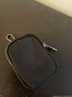 Photo of the front side of a small black fabric change pouch with a metal clip at the corner and a metal zipper around the edge.