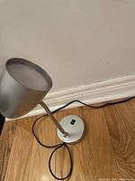 Desk lamp with silver shade, flexible neck, round base with switch, and black power cord on wood floor.