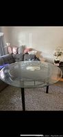 Round clear glass dining table with black legs in a living room setting.