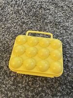 Yellow plastic egg holder tray displayed on a carpeted surface with twelve dome-shaped compartments and a handle.