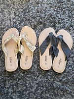 Two pairs of women's size 6 flip flops on a gray carpet, one pair with gold metallic fabric straps and the other pair with dark gray fabric bow straps.