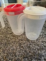Photo of two clear plastic juice jugs with handles, one red lid and one white lid, placed on carpet.