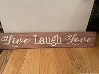 Rectangular wooden wall sign with the phrase 'Live Laugh Love' written in white letters on a brown rustic background, placed on a wooden floor against a white wall.