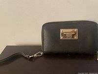 Front view of the black leather Michael Kors wallet with gold logo plaque and wristlet strap on a dark surface against a beige wall.