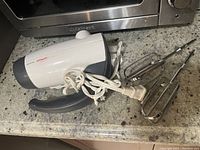 Side view of the Sunbeam hand mixer with two detachable chrome beaters laid beside it on granite countertop.