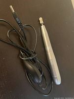 Photo showing silver metal nail drill handpiece connected to a black power cord with inline control on dark surface.