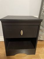 Front view of the small black wooden nightstand showing a closed drawer with a silver oval knob and an open shelf below it.