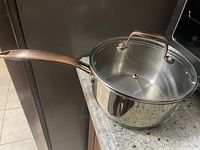 Cooking pot placed on granite countertop, showing reflective stainless steel body, copper-colored handle, and glass lid with steam vent.