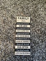 Rectangular wooden hanging sign with painted black and white text listing family rules on a carpet background.