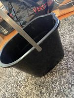 Photo shows a small, black, oval-shaped plastic bucket with a metal handle resting on a carpeted floor.