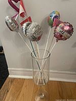 Tall clear glass vase with six decorative small foil balloons on white sticks featuring celebration messages.