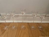 Five clear glass tea light candle holders with varying stem heights displayed against a wall on wooden floor.