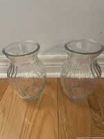 Photo showing a set of two clear glass vases with ribbed pattern and flared open tops on a wooden floor against a white wall.
