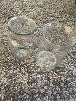 Two clear glass vases placed on a textured carpet, one with a bulging body and flared top, the other cylindrical with wide open top.