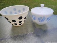 Photo of two vintage white bowls placed outdoors on a glass surface. One bowl has black polka dots and is open-topped. The other bowl has blue floral patterns, a lid with knob, and is oven safe.