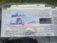 Box showing the Rack-Pack rooftop cargo bag mounted on a red SUV roof, product branding and usage instructions visible on box.