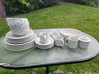 Photo of the ceramic dish set arranged outdoors on a glass patio table. Includes stacked bowls, plates, mugs, small plates, and a small lidded jug or sugar bowl, all with a white base and black heart design.