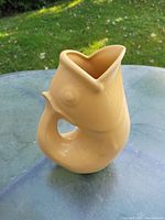 Photo of a cream-colored ceramic fish-shaped jug with curled fish tail handle on a glass table outdoors with grass background.