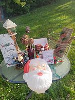 Photo showing an outdoor round glass table holding various Christmas decorations including a large light-up Santa face, Merry and Bright flag on wooden stake, rustic wooden sled with reindeer decoration, brown vase with twig decorations, assorted baubles and small holiday figures.