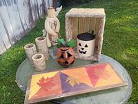 Photo showing various outdoor fall decor items on glass table including a wooden crate, pumpkin container, wooden figurine, three votive holders, and a framed leaf art