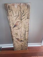 Vertical wooden wall art panel featuring two robins and foliage painted on a distressed surface, resting against a wall.