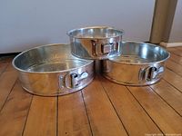 Photo of three metal round spring-form pans with latch locks placed on wooden floor.