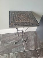 Photo of a metal side/accent table with a square decorative top featuring intricate embossed floral and geometric patterns. The table has curved metal legs and is suitable for placing in a corner of a room.
